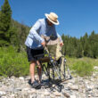 Man begins setting up a tan Adjustable Rocker chair on a rocky riverside trail.