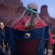 Woman in a red jacket and cap sitting in a navy topo Firepit Rocker by a campfire.