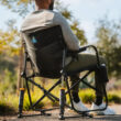 A person relaxing outdoors in a black Freestyle Rocker Elite chair with a mesh back, cup holder, and sturdy frame.